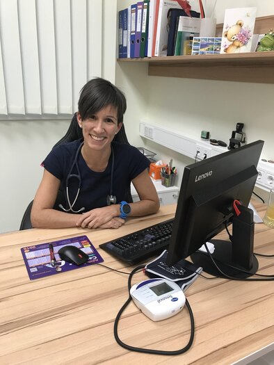 Eine lächelnde Ärztin sitzt an ihrem Schreibtisch in einer Praxis. Der Tisch ist mit einem Computer, einem Tastatur und medizinischen Geräten ausgestattet. | © Dr. Thier | Arztpraxis