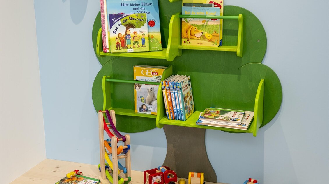 Ein kindgerechter Spielbereich mit einem Bücherregal in Form eines Baumes. Es sind verschiedene Bücher und Spielzeuge auf einem Holzregal angeordnet. | © Lorenz Masser