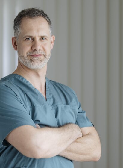 Ein Arzt in grüner Arbeitskleidung steht mit verschränkten Armen. Er blickt selbstbewusst in die Kamera.