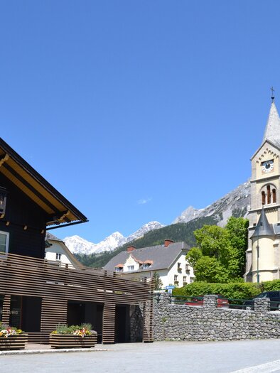 A picturesque village with a traditional wooden house and an impressive church in the background. The mountains are clearly visible, and the sky is bright blue. | © tita.at