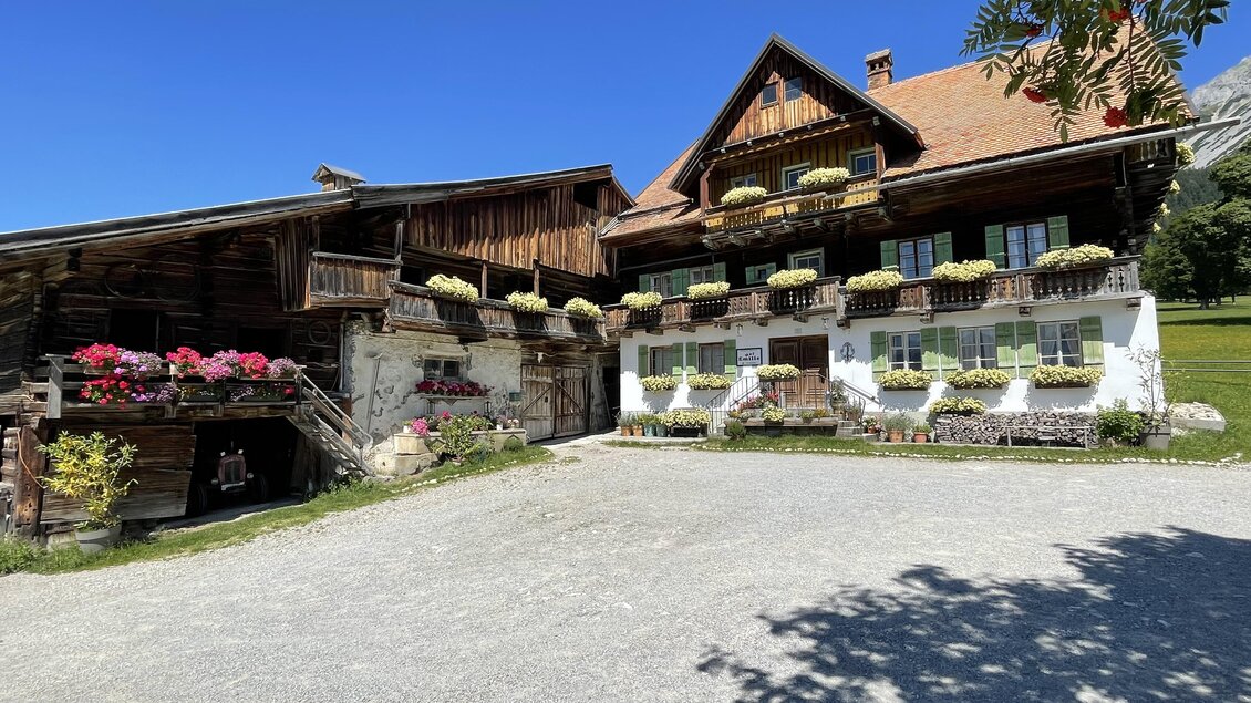 Ein traditionelles Bauernhaus mit Holzfassade und blühenden Balkonkästen. Umgeben von einem grünen Rasen und klarem blauen Himmel. | © photo-austria.at/Hans-Peter Steiner