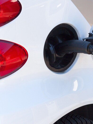 A white electric car with a charging cable connected to a charging station. In the background, the taillights of the vehicle are visible. | © Symbolfoto