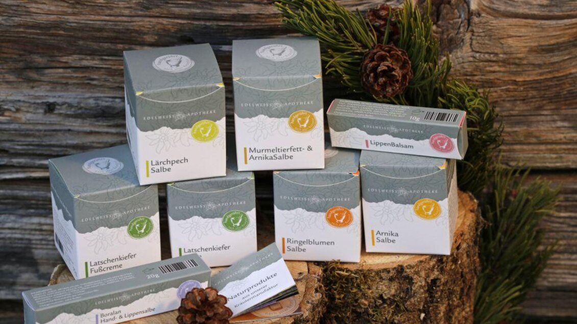 Eine Sammlung von Heilungsprodukten in stilvollen Verpackungen auf einem Holzstamm. Im Hintergrund sind grüne Pflanzen und rustikales Holz zu sehen. | © Edelweiss Apotheke