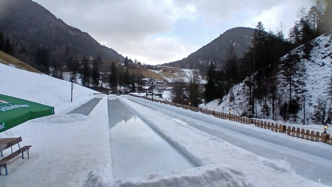Eine schneebedeckte Landschaft mit einer Rodelbahn, die durch die Berge verläuft. Im Hintergrund sind bewaldete Hügel und ein klarer Himmel sichtbar. | © Almstube Schlattinger