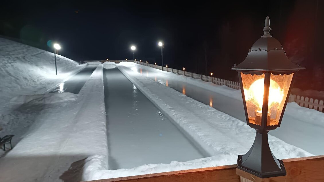 Eine schneebedeckte Rodelbahn bei Nacht mit hellen Lichtern. Im Vordergrund steht eine Laterne, die warmes Licht ausstrahlt. | © Almstube Schlattinger