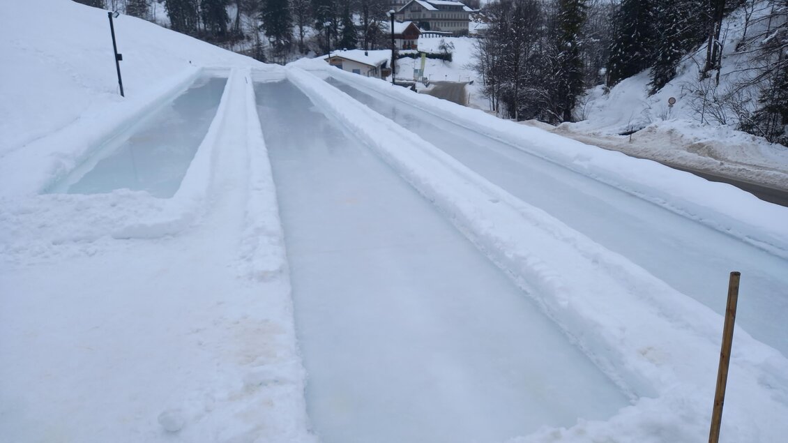 Eine verschneite Landschaft mit zwei Eislaufbahnen. Im Hintergrund sind Bäume und kleine Gebäude sichtbar. | © Almstube Schlattinger