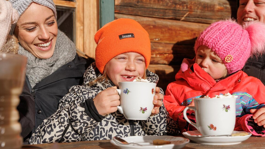 Eine fröhliche Familie sitzt zusammen vor einer Holzfassade und genießt heiße Schokolade aus großen Tassen. Kinder in bunten Winterjacken lächeln und haben Spaß. | © Waldhäuslalm/Huber Martin 