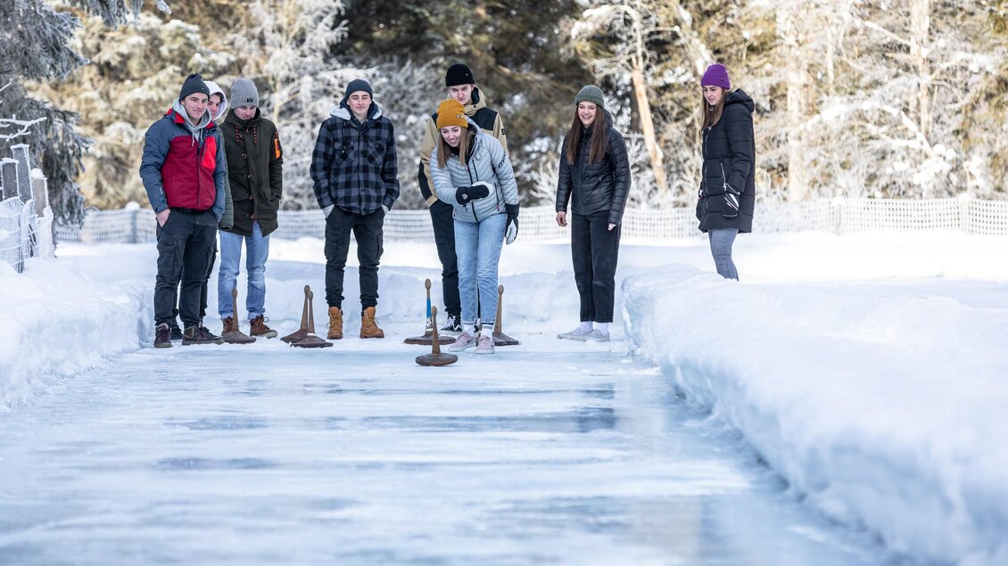 Eine Gruppe von sechs Personen steht auf einer gefrorenen Oberfläche im Winter. Sie spielen eine Figur auf dem Eis und genießen die kalte, verschneite Umgebung. | © Waldhäuslalm/Martin Huber