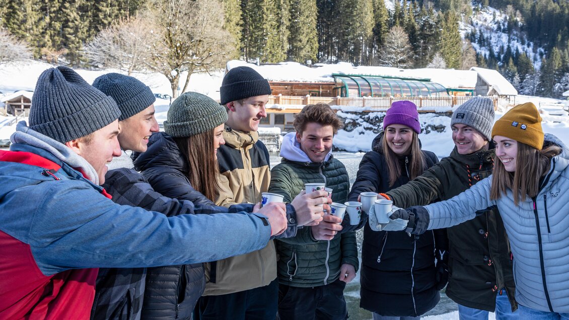 Eine Gruppe von acht Personen steht im Schnee und stößt mit Getränken an. Im Hintergrund sind schneebedeckte Bäume und eine Hütte zu sehen. | © Waldhäuslalm/Martin Huber