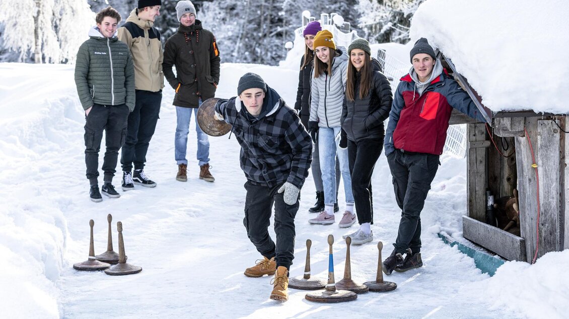 Eine Gruppe von Menschen genießt eine winterliche Aktivität im Schnee. Jemand spielt eine Art Curling auf einer eisigen Bahn, während die anderen zuschauen. | © Waldhäuslalm/Martin Huber