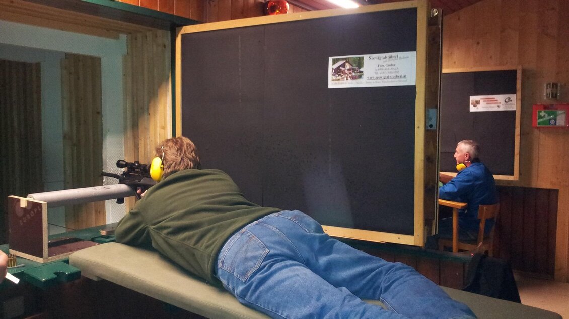 Eine Person liegt auf einer Schießbank und zielt mit einem Gewehr. Im Hintergrund sitzt eine weitere Person und beobachtet. | © Erzherzog-Johann-Schießstätte Aich