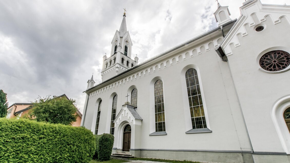 Eine weiße Kirche mit einem hohen Turm und großen Fenstern. Im Vordergrund befinden sich grüne Büsche und ein bewölkter Himmel. | © Gerhard Pilz