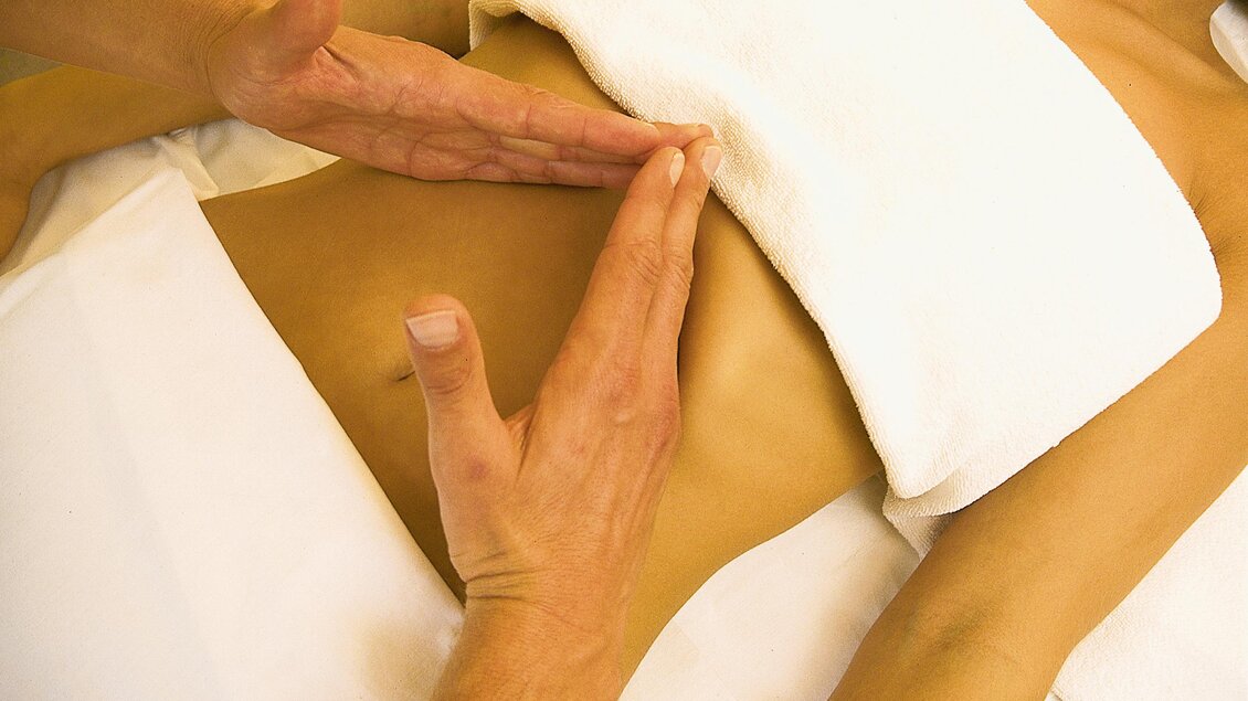 Eine Person erhält eine Massage mit Händen, die sanft über den Bauchbereich gleiten. Ein Handtuch ist auf der Haut platziert, um eine entspannende Atmosphäre zu schaffen. | © Hotel Spanberger
