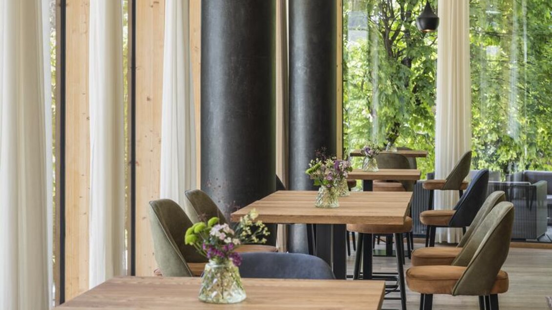 Ein modernes Café mit Holztischen und stilvollen Stühlen. Große Fenster lassen viel Licht herein und bieten einen Blick ins Grüne. | © Falkensteiner Hotel Schladming/ Fotograf Walter Luttenberger