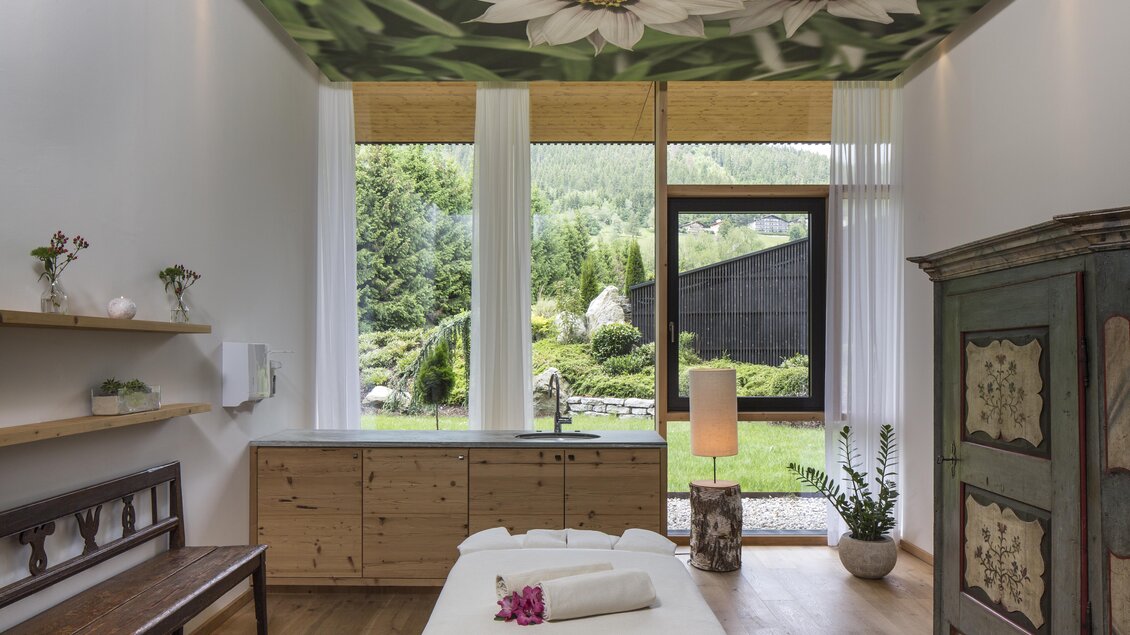 Ein helles Zimmer mit großen Fenstern, die auf einen grünen Garten blicken. Die Wände sind mit einer schönen Blumendekoration und einer rustikalen Holzmöbel ausgestattet. | © Falkensteiner Hotel Schladming/ Fotograf Walter Luttenberger