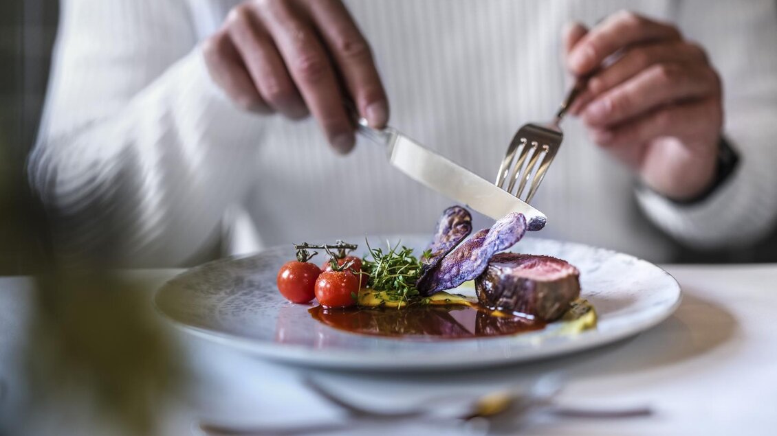Ein elegantes Gericht auf einem Teller mit zarten Stückchen Fleisch, bunten Kartoffeln und Kirschtomaten. Eine Hand schneidet sorgfältig das Essen mit einem Messer und einer Gabel. | © Falkensteiner Hotels und Residences