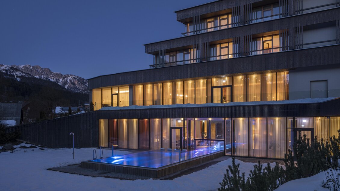Ein modernes Hotel mit großen Fenstern und einem beleuchteten Außenbereich. Der Pool ist von Schnee umgeben und die Berge sind im Hintergrund sichtbar. | © Falkensteiner Hotel Schladming/ Fotograf Walter Luttenberger