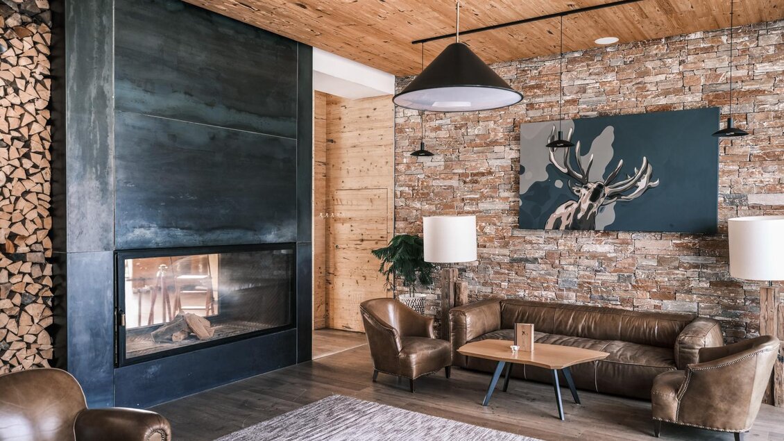 Ein modernes Wohnzimmer mit einem gemütlichen Kamin und einer Steinwand. Die Einrichtung besteht aus braunen Ledermöbeln und stilvollen Lampen. | © Falkensteiner Hotel Schladming/ Fotograf Dragan Dokmanovic