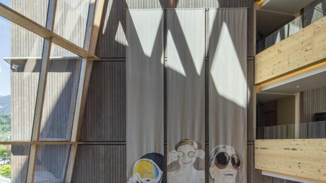 Eine moderne Hotellobby mit großen Fenstern und Holzelementen. An der Wand hängen dekorative Vorhänge mit Illustrationen von Gesichtern. | © Falkensteiner Hotel Schladming/ Fotograf Walter Luttenberger