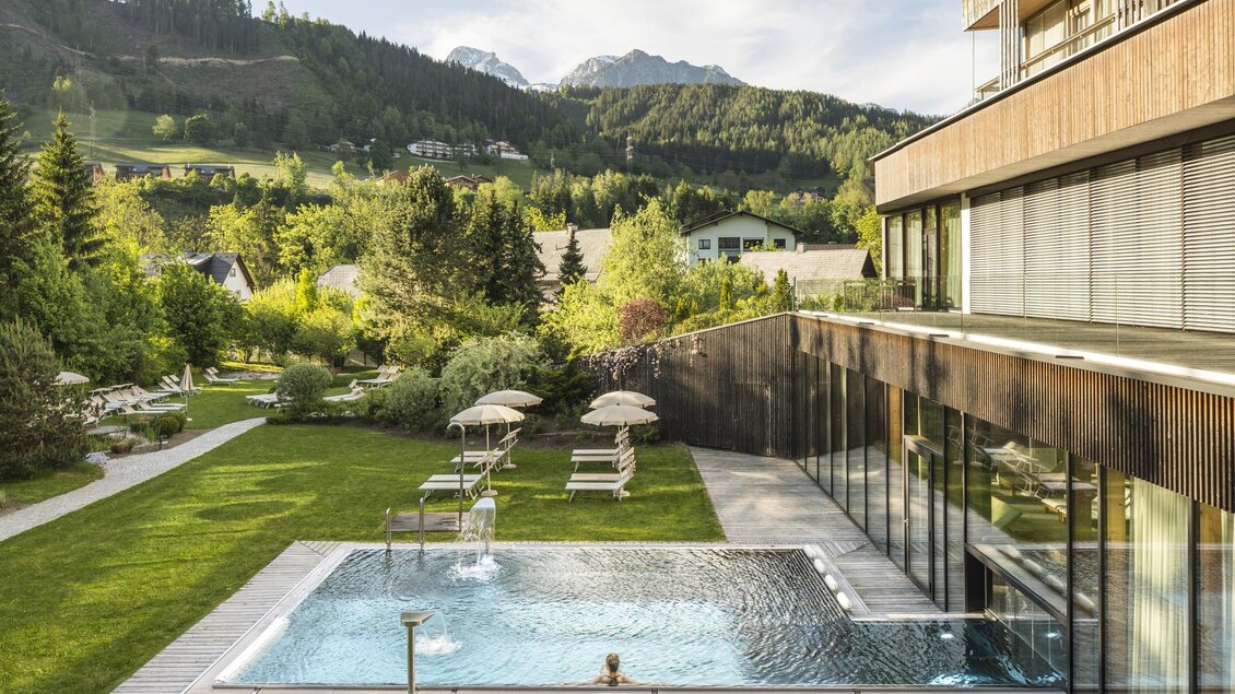 Ein modernes Hotel mit einem Swimmingpool und einer schönen Gartenlandschaft. Im Hintergrund sind bewaldete Berge und ein klarer Himmel zu sehen. | © Falkensteiner Hotel Schladming/ Fotograf Walter Luttenberger