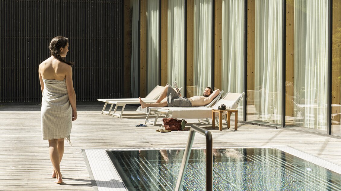 Eine Frau in einem Handtuch geht am Pool vorbei, während ein Mann auf einer Liege entspannt. Die Umgebung ist modern und hell mit großen Fenstern. | © Falkensteiner Hotel Schladming/ Fotograf Walter Luttenberger