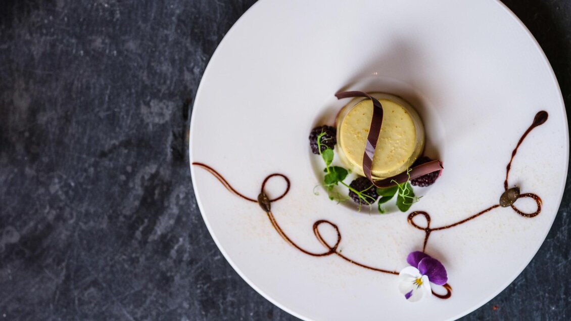 Ein elegantes Dessert auf einem weißen Teller, dekoriert mit Schokoladenschaifen und frischen Beeren. Dazu wachsen zarte Pflanzen und eine kleine Blume, die das Gericht ansprechend präsentiert. | © Falkensteiner Hotel Schladming/ Fotograf Dragan Dokmanovic