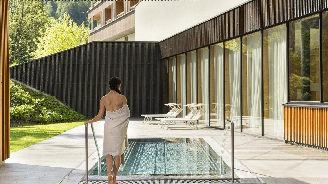 Eine Person steht am Rand eines Innenpools und schaut nach draußen. Der Bereich ist modern gestaltet, umgeben von großen Fenstern und einer grünen Landschaft. | © Falkensteiner Hotel Schladming/ Fotograf Walter Luttenberger
