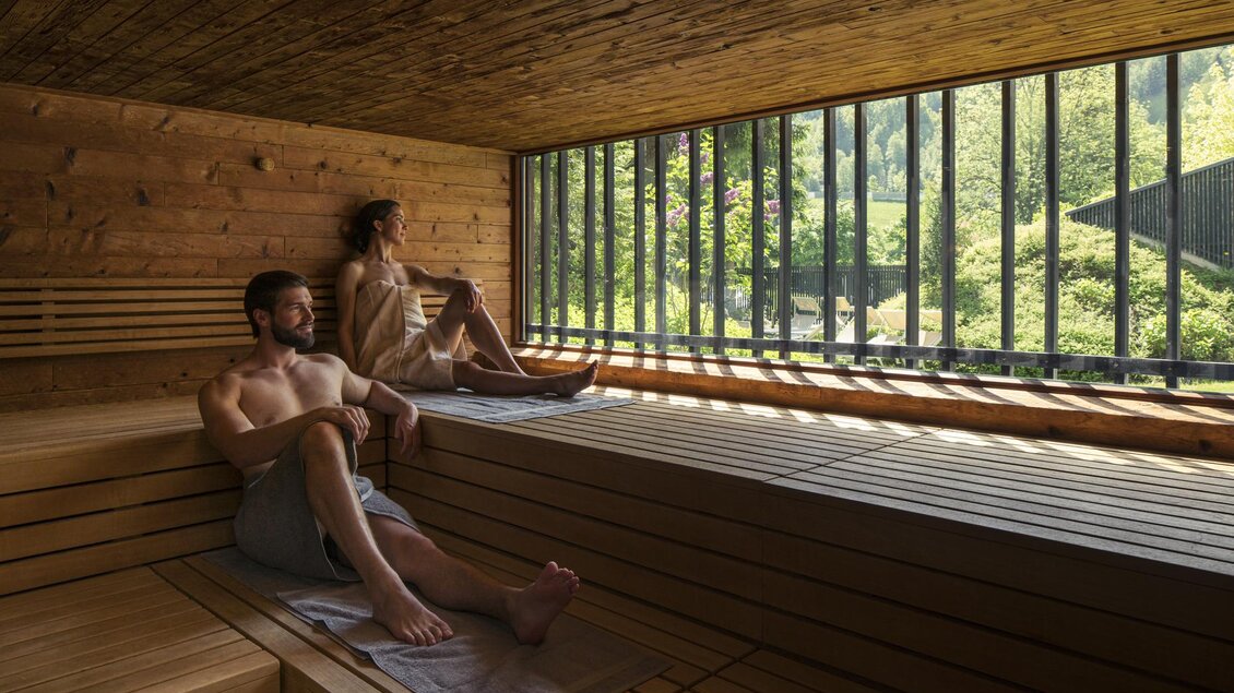 Zwei Personen entspannen in einer Holzsauna mit großen Fenstern. Draußen sieht man eine grüne Landschaft. | © Falkensteiner Hotel Schladming/ Fotograf Walter Luttenberger