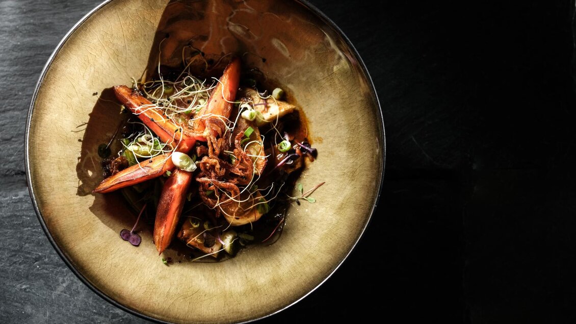 Ein kunstvoll angerichtetes Gericht in einer Schüssel. Die Speise besteht aus Karotten, Kräutern und einer dunklen Sauce. | © Falkensteiner Hotel Schladming/ Fotograf Dragan Dokmanovic