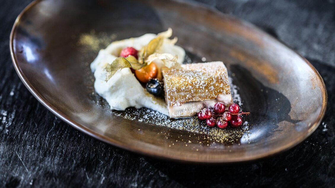 Ein elegantes Dessert auf einem braunen Teller, bestehend aus einer süßen Füllung, Sahne und frischen Beeren. Das Gericht ist kunstvoll dekoriert und sieht sehr ansprechend aus. | © Falkensteiner Hotel Schladming/ Fotograf Dragan Dokmanovic