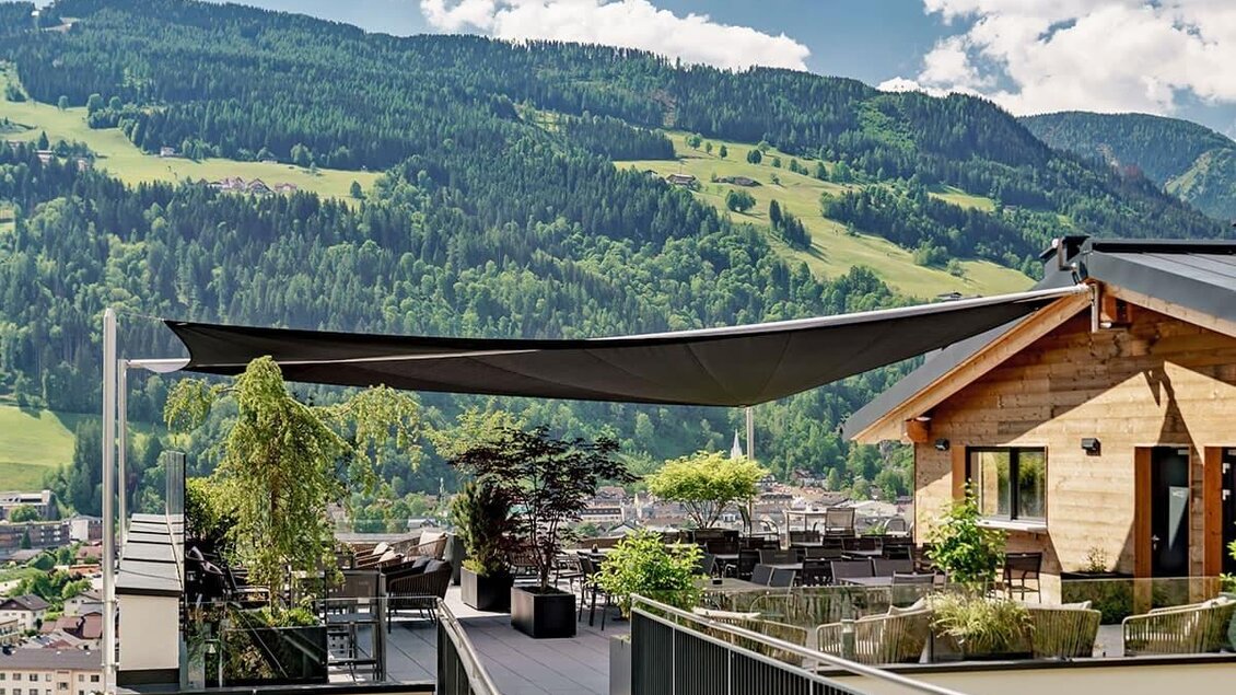 Eine moderne Terrasse mit einem Sonnensegel und Blick auf grüne Berge. Die Umgebung ist mit Pflanzen und einem Holzgebäude gestaltet. | © Aparthotel Ferienalm