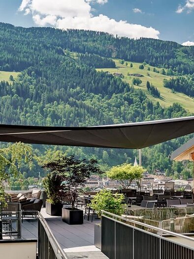 Eine moderne Terrasse mit einem Sonnensegel und Blick auf grüne Berge. Die Umgebung ist mit Pflanzen und einem Holzgebäude gestaltet. | © Aparthotel Ferienalm