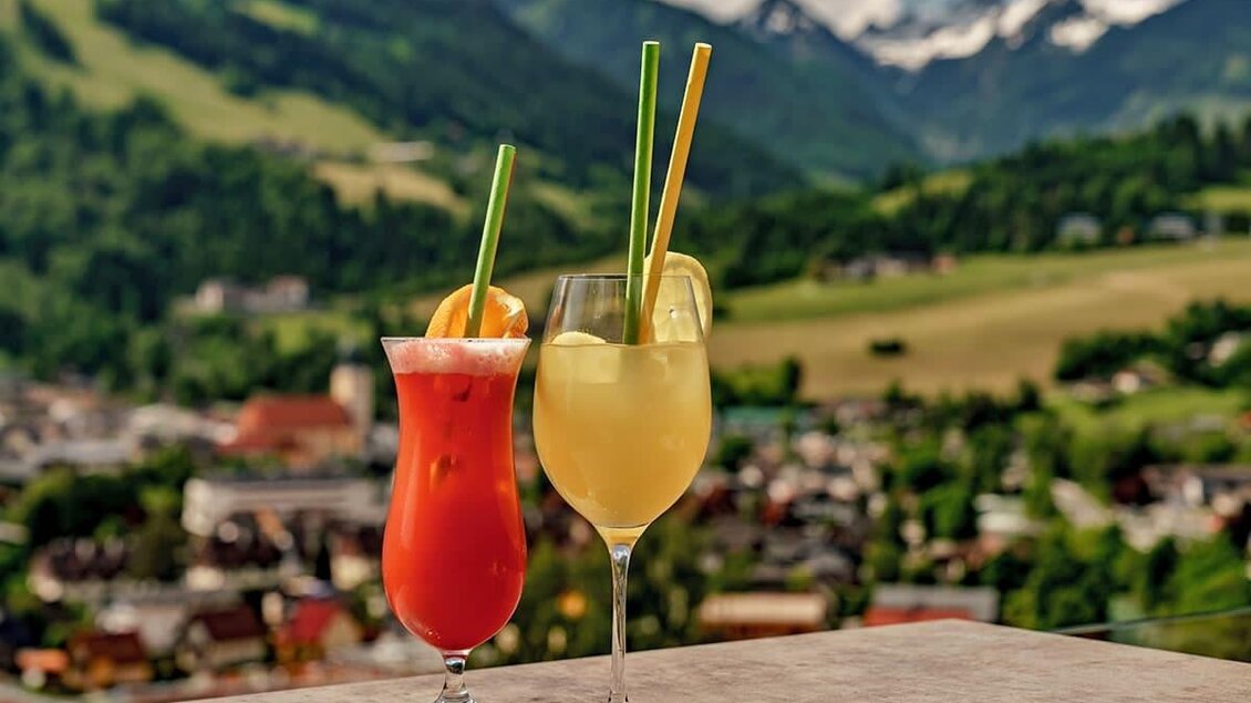 Zwei erfrischende Cocktails stehen auf einem Tisch mit Blick auf eine malerische Berglandschaft. Im Hintergrund sind grüne Hügel und ein blauer Himmel mit Wolken zu sehen. | © Aparthotel Ferienalm
