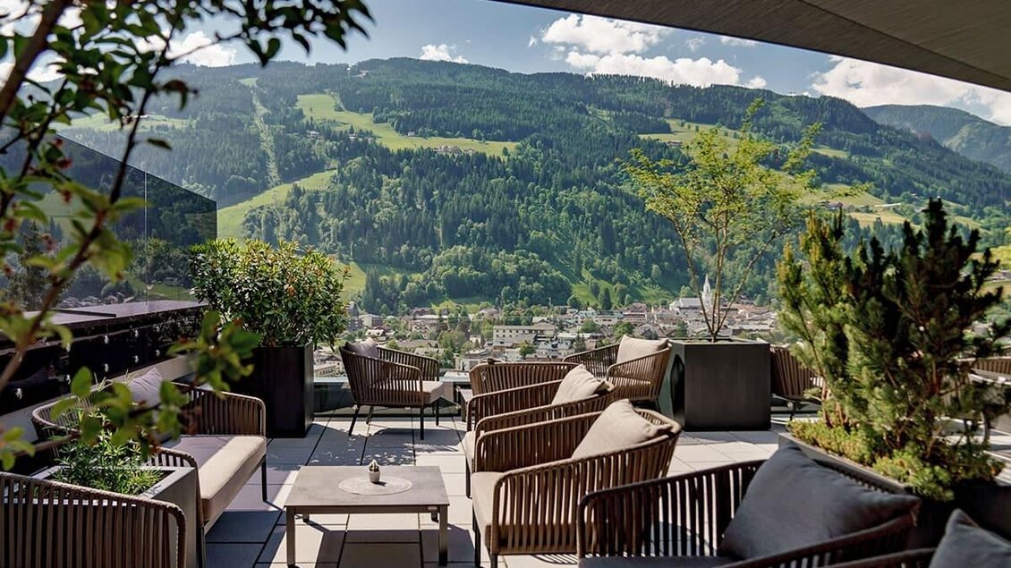 Eine moderne Terrasse mit bequemen Möbeln und Pflanzen. Im Hintergrund sind grüne Berge und ein klarer Himmel zu sehen. | © Aparthotel Ferienalm
