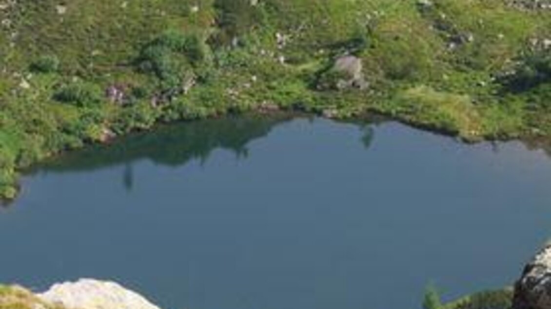 Ein ruhiger Bergsee umgeben von sanften Hügeln und grüner Vegetation. Im Vordergrund fließt ein kleiner Wasserfall in den See. | © Fischen im Sattental
