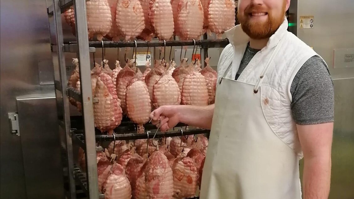 Ein lächelnder Mann in einer weißen Schürze steht neben einem Regal mit geräuchertem Fleisch. Im Hintergrund sind Metalltüren zu sehen. | © Fleischhauerei Tasch