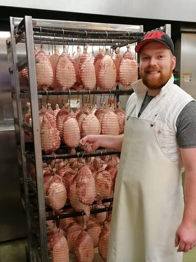 Ein lächelnder Mann in einer weißen Schürze steht neben einem Regal mit geräuchertem Fleisch. Im Hintergrund sind Metalltüren zu sehen. | © Fleischhauerei Tasch