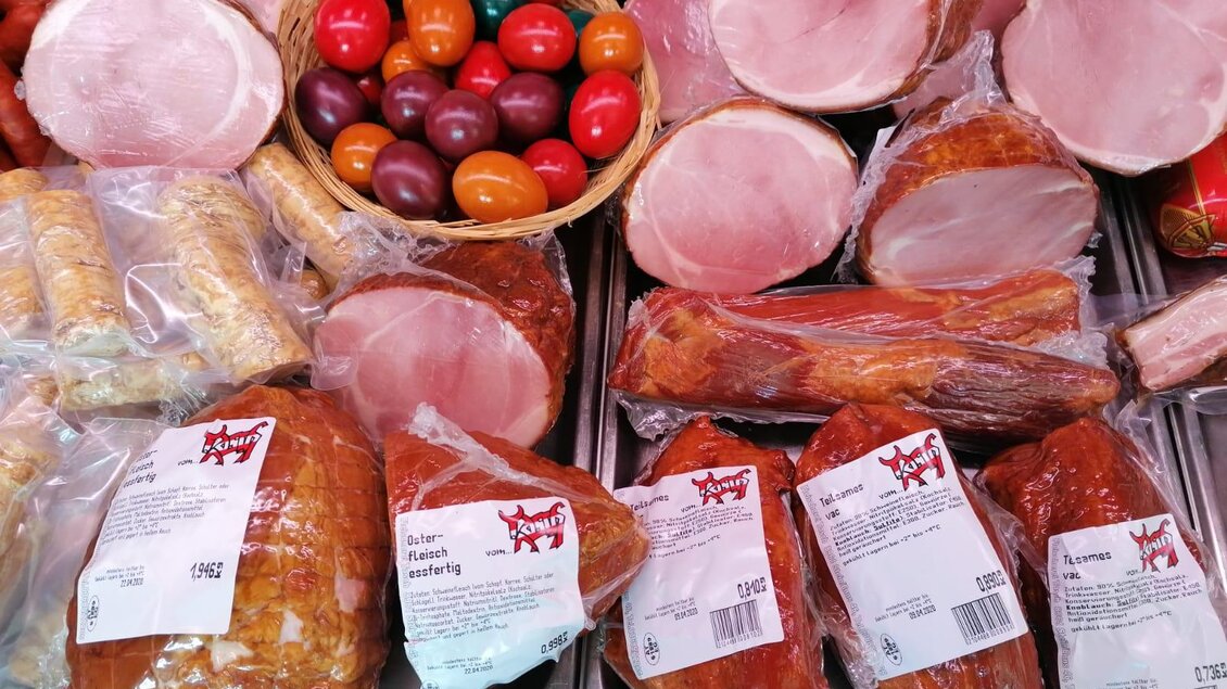 Eine Auswahl an verschiedenen Wurst- und Schinkenprodukten liegt auf einem Tisch. Daneben befinden sich bunte, österliche Dekorations-Eier in einem Korb. | © Fleischhauerei Tasch