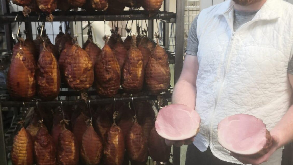 Ein Mann steht neben einem Regal mit geräuchertem Schinken. Er hält zwei Schalen mit Schinken in der Hand. | © Fleischhauerei Tasch