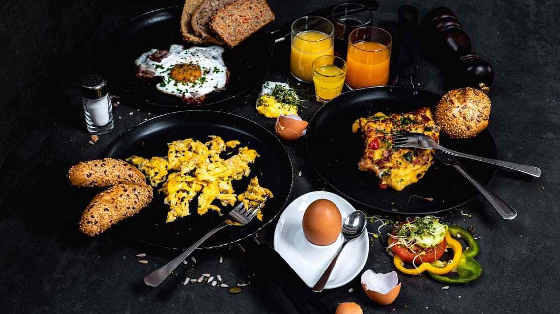 Ein reichhaltiges Frühstück mit Rührei, Brot, frischem Gemüse und verschiedenen Getränken. Die Mahlzeit ist ansprechend auf einem dunklen Tisch angerichtet. | © Stefan Trocha