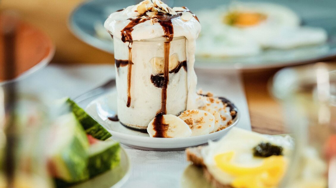 Ein köstliches Dessert in einem Glas mit Vanilleeis, Schokolade und Banane. Im Hintergrund sind frisches Obst und ein einfaches Frühstück zu sehen. | © Stefan Trocha