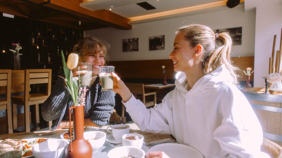 Zwei Frauen stoßen in einem Café mit Getränken an. Auf dem Tisch stehen verschiedene Frühstücksgerichte. | © Stadthotel brunner