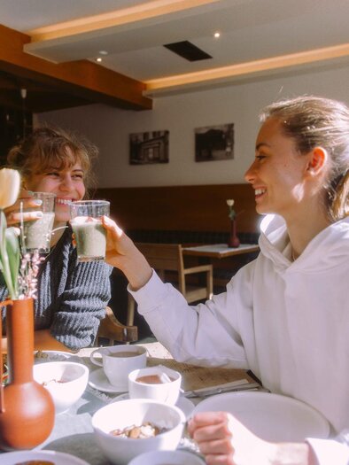 Two women toast with drinks in a café. Various breakfast dishes are on the table. | © Stadthotel brunner