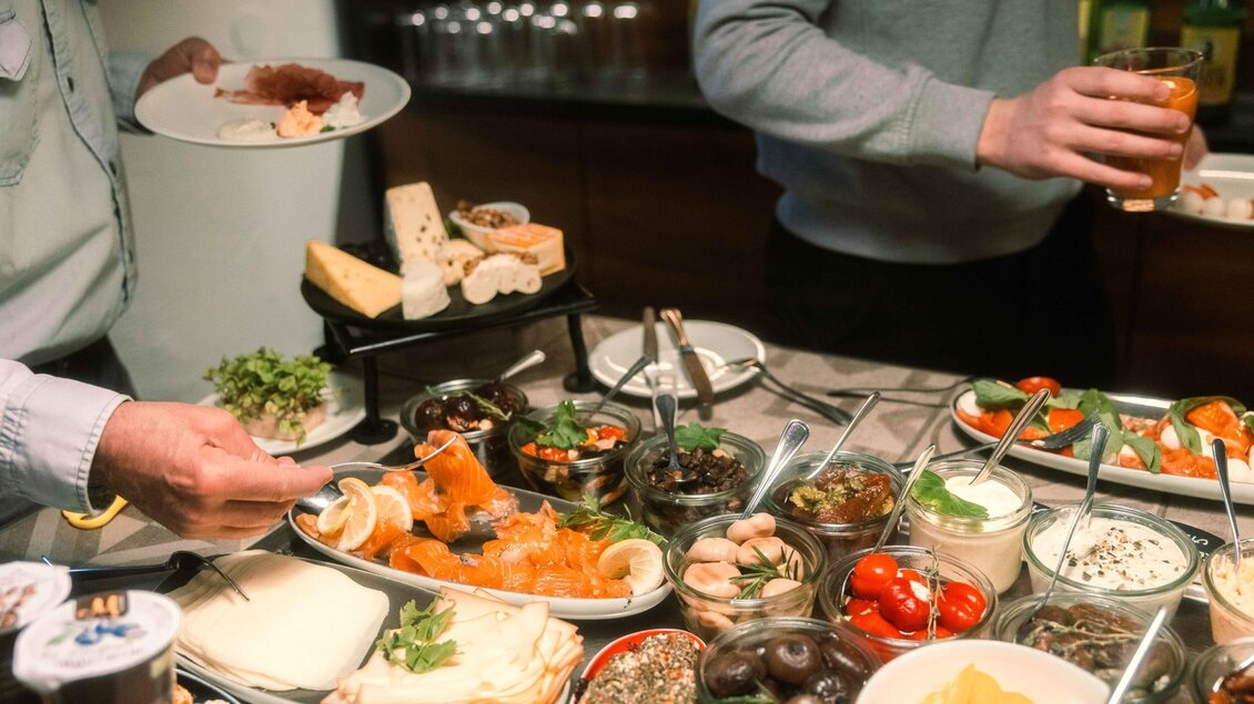 Eine reichhaltige Auswahl an Speisen auf einem Buffet, darunter Lachs, Käse, Oliven und frisches Gemüse. Zwei Personen sind mit Tellern und Getränken beschäftigt. | © Stadthotel brunner