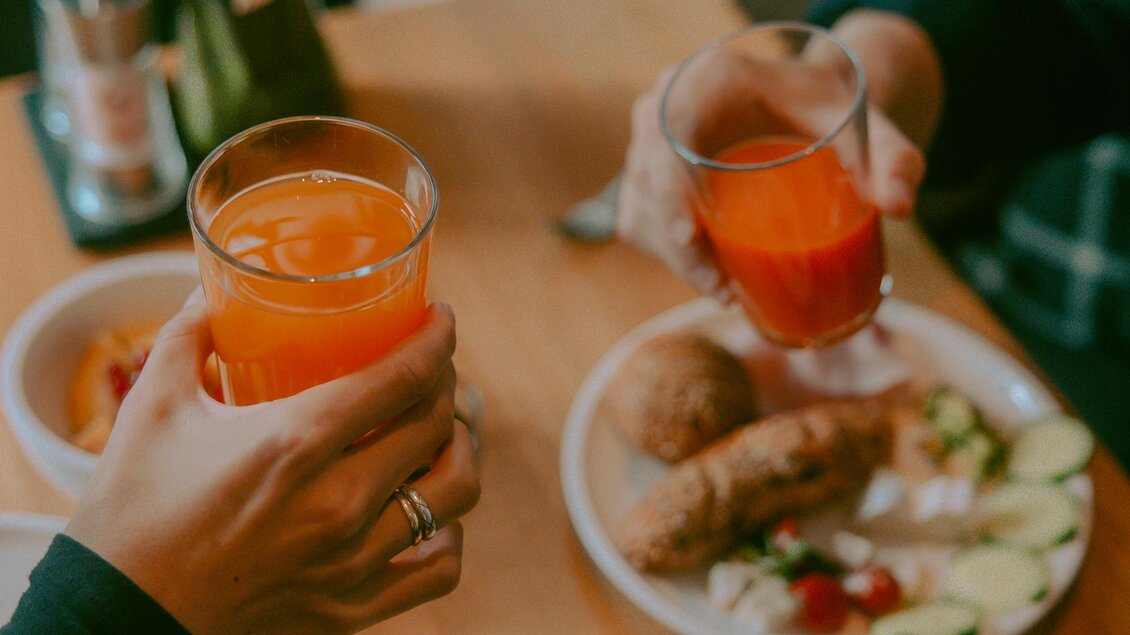 Ein gemütliches Frühstück mit frischen Brötchen und Gemüse auf dem Tisch. Zwei Personen stoßen mit Gläsern voller Saft an. | © Stadthotel brunner