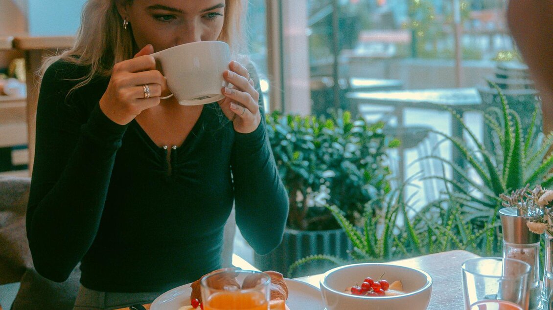 Eine Frau trinkt aus einer Tasse, während sie an einem Tisch sitzt. Auf dem Tisch sind verschiedene Teller mit Essen und ein Glas Saft zu sehen. | © Stadthotel brunner