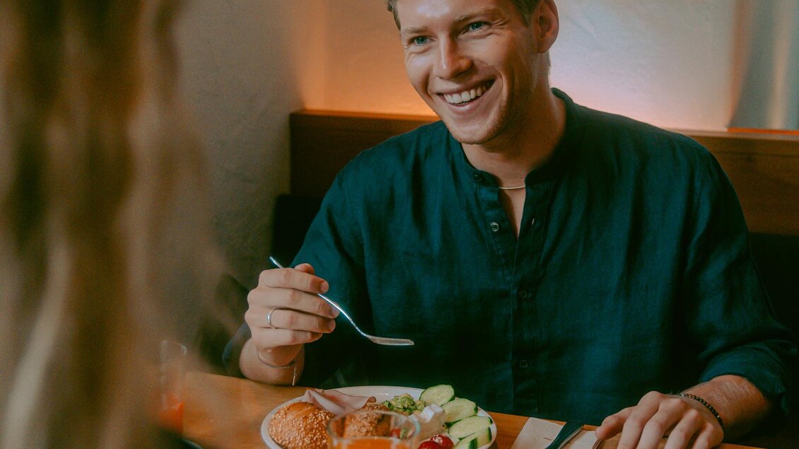Ein Mann sitzt lächelnd an einem Tisch und genießt eine Mahlzeit. Auf dem Tisch sind zwei Teller mit Essen und Getränke zu sehen. | © Stadthotel brunner