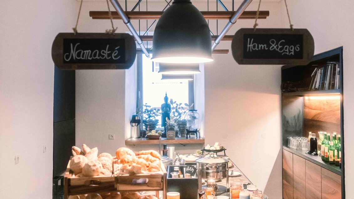 Ein einladendes Frühstücksbuffet mit frisch gebackenem Brot und verschiedenen Köstlichkeiten. An der Wand hängen zwei Schilder mit den Aufschriften „Namasté“ und „Ham & Eggs“. | © Stadthotel brunner