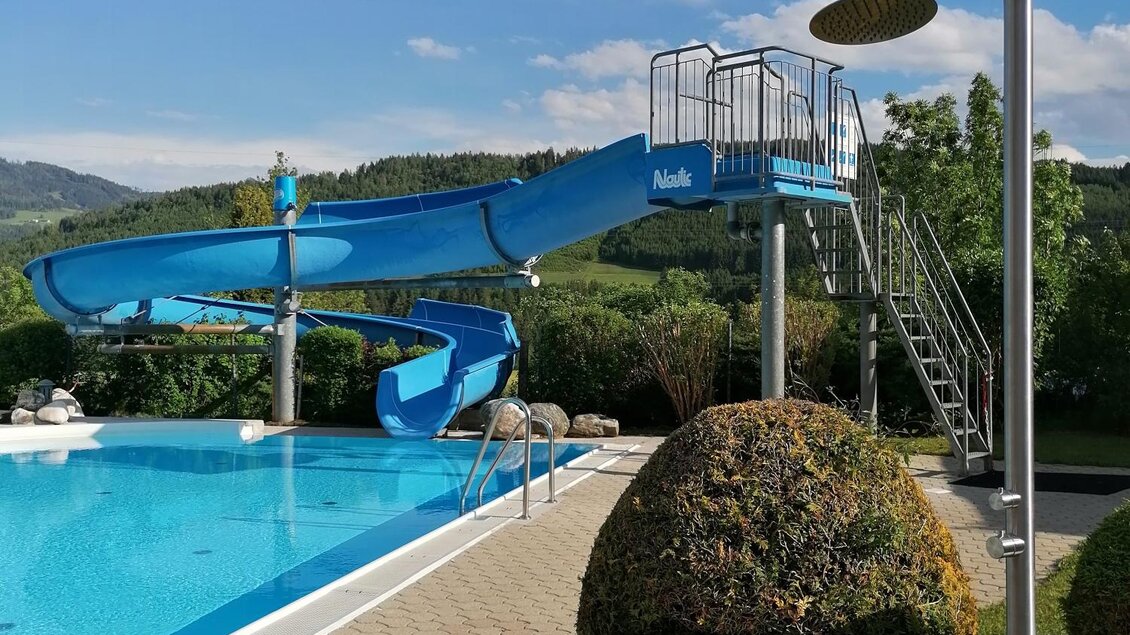 Ein Schwimmbad mit einer langen Wasserrutsche und einem Duschturm. Die Umgebung ist grün und einladend mit schönem Himmel. | © Gemeinde Mitterberg-Sankt Martin