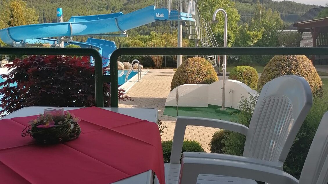 Ein gemütlicher Balkon mit einem Tisch und Stühlen. Im Hintergrund sind ein Schwimmbad und eine Rutsche zu sehen, umgeben von grünen Hügeln. | © Gemeinde Mitterberg-Sankt Martin